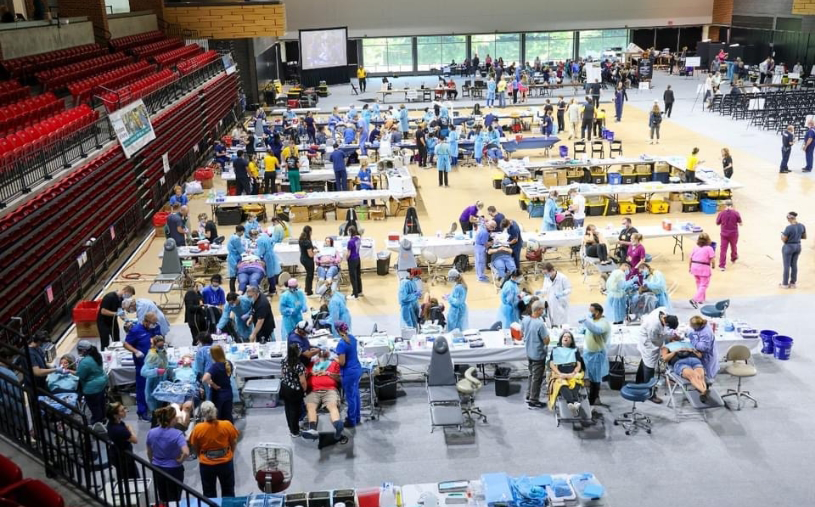 Several dental care stations set up in a gym with dozens of people receiving dental treatment